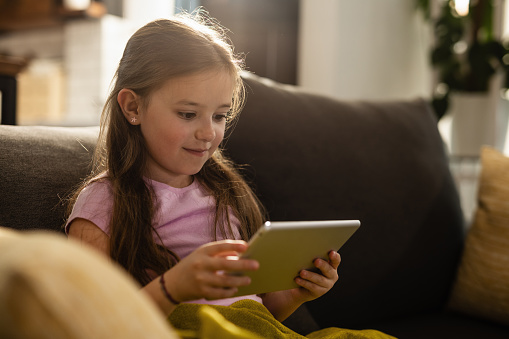 Smiling little girl reading on touchpad while sitting on the sofa and relaxing in the living room